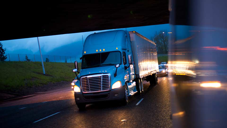 Camión de transporte de carga circulando por una carretera durante la noche, con las luces encendidas, destacando la seguridad en el transporte en condiciones de baja visibilidad.
