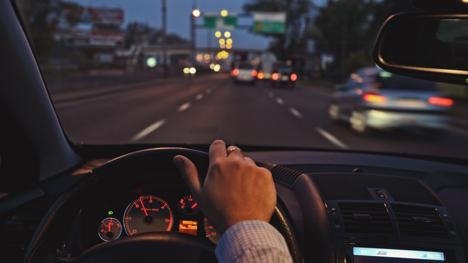 Vista desde el interior de un vehículo circulando por una avenida al anochecer, con la mano del conductor sobre el volante y varios autos al frente. La imagen representa la importancia de mantener la atención en el camino y prevenir riesgos al manejar.