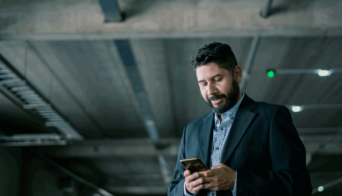 Hombre de negocios con barba usando el celular en un estacionamiento techado, vestido con saco oscuro y camisa estampada, concentrado en la pantalla de su teléfono.