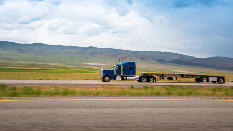 Camión de carga azul con remolque vacío en carretera montañosa bajo un cielo parcialmente nublado, representando la logística de transporte.