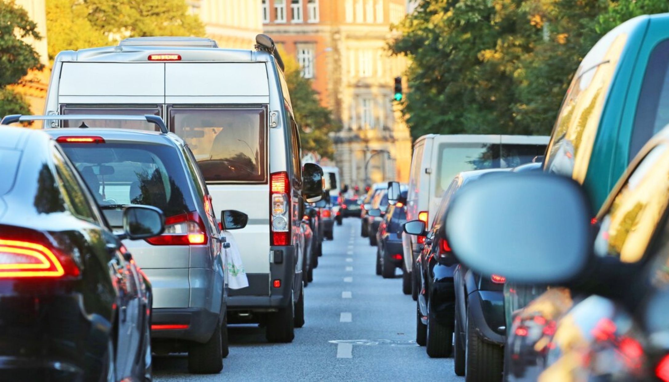 Verkehrsstau auf einer Stadtstraße, Blick auf eine Fahrspur mit Stoßstange-an-Stoßstange-Autos, darunter ein SUV und ein weißer Lieferwagen. Im Hintergrund säumen Gebäude und Bäume die Straße, ein grünes Verkehrssignal ist sichtbar. Die Beleuchtung deutet auf den späten Nachmittag hin.