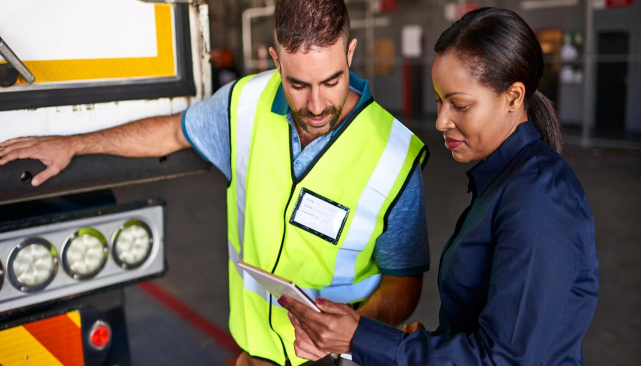 Vrachtwagenchauffeur in geel hesje en vrouwelijke wagenparkbeheerder bespreken informatie op een tablet bij een vrachtwagen.